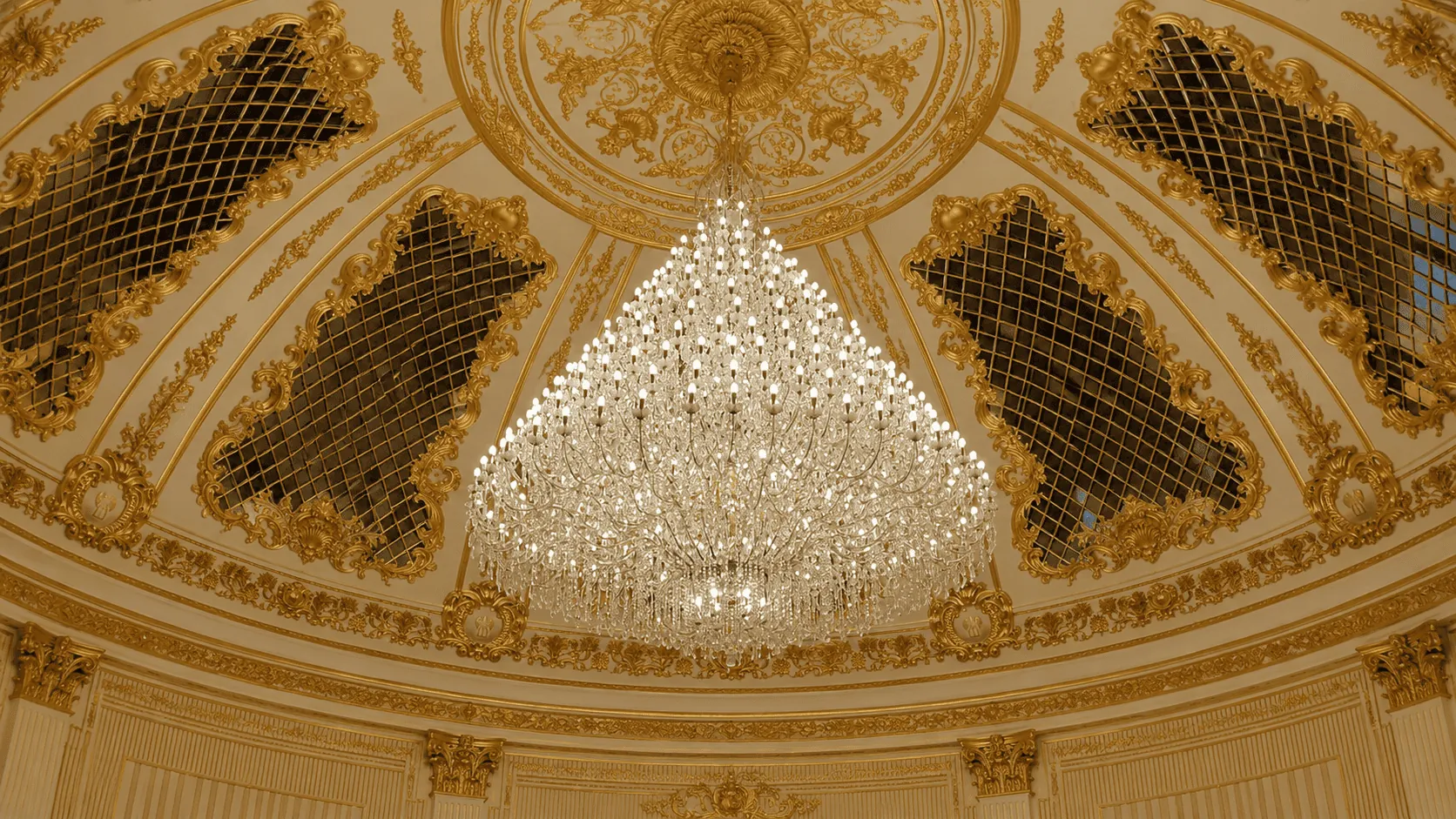 Lampadario monumentale in cristallo sospeso sotto la cupola dorata di una sala storica.