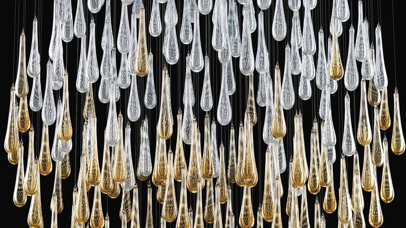 Una cascata di gocce in vetro soffiato, trasparenti e in foglia oro, su fondo nero.