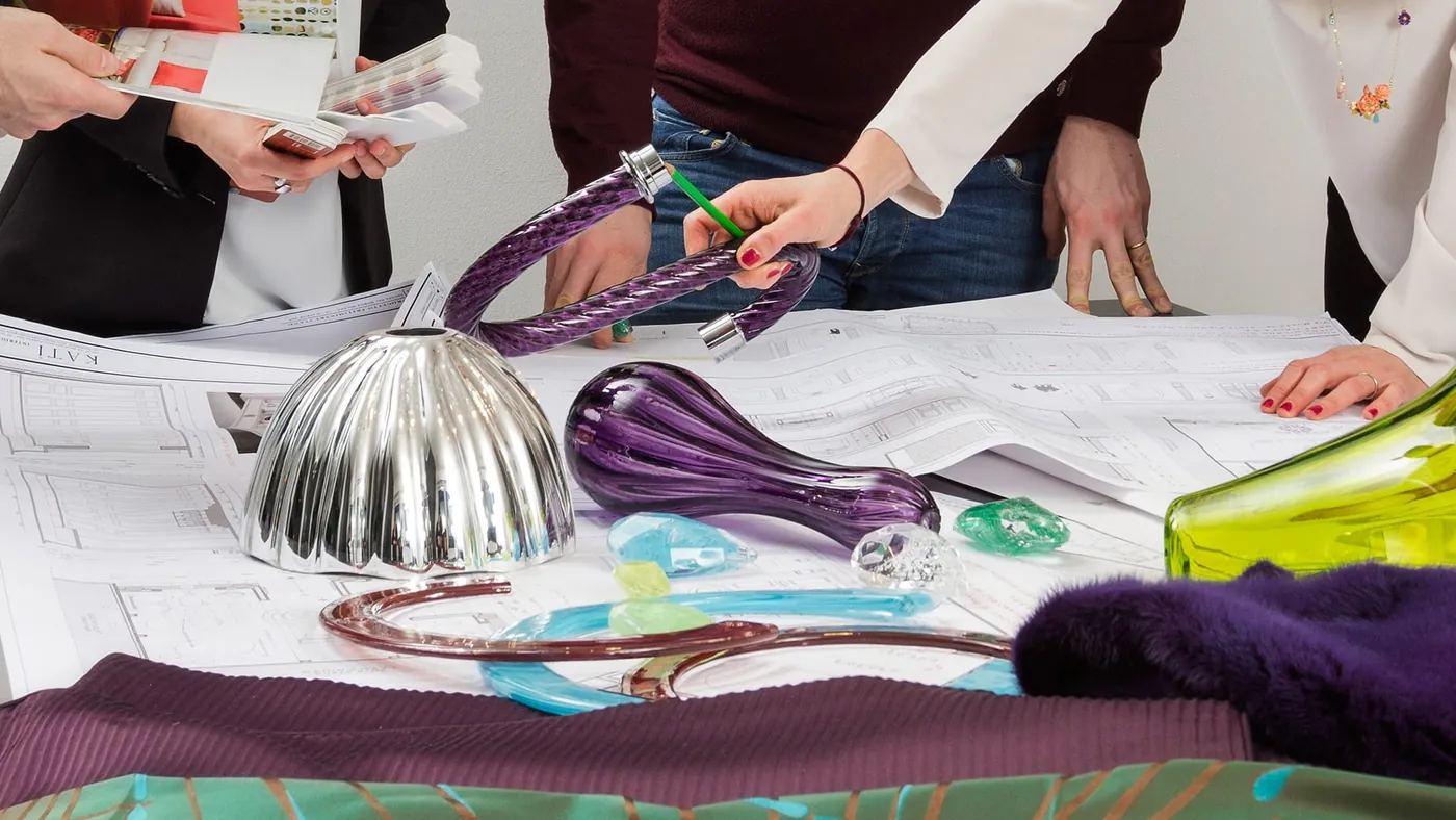 Tavolo di progetto: disegni esecutivi, campioni di vetro di Murano viola e verde, tessuti, e le mani che li confrontano.