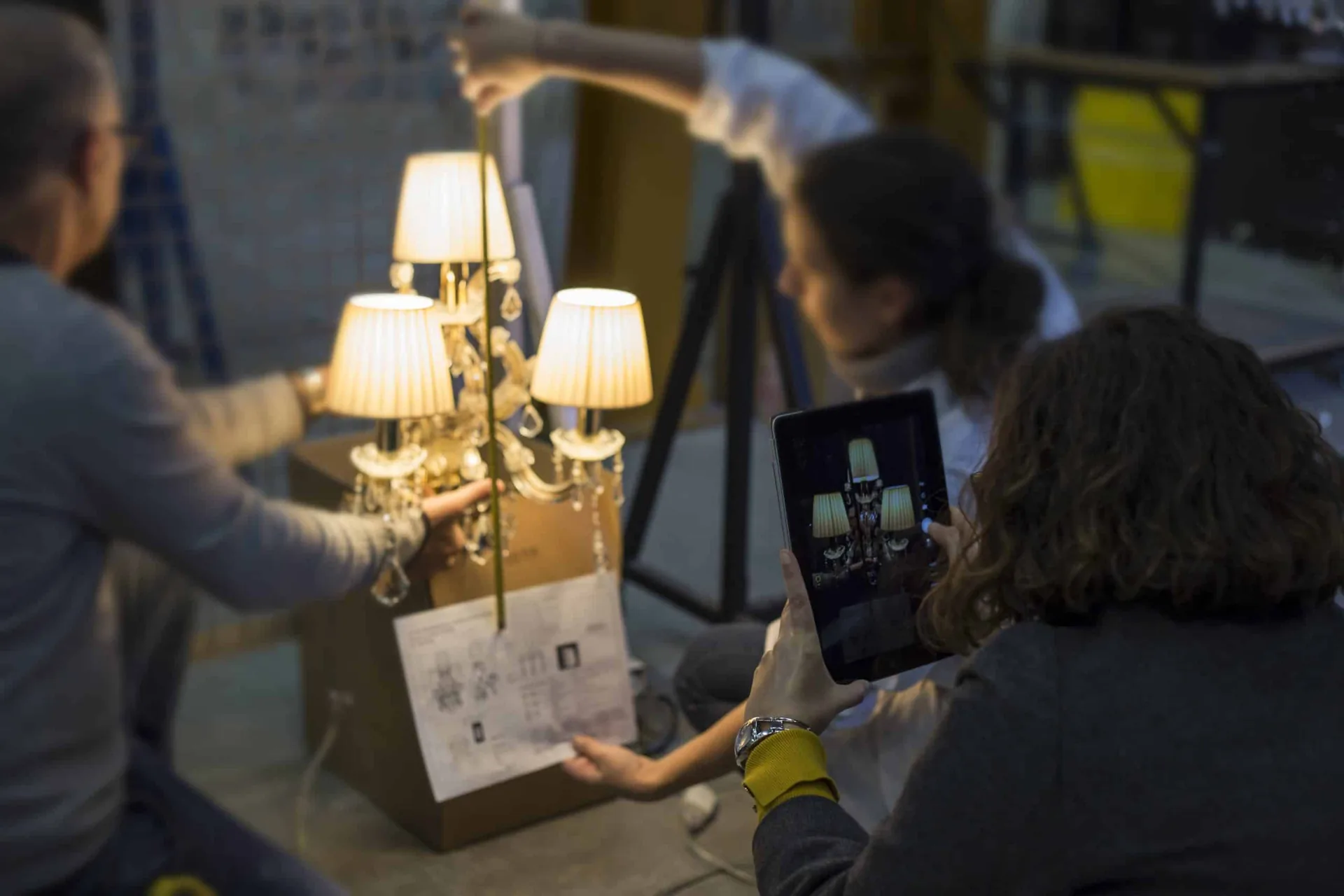 Due persone verificano un lampadario acceso confrontandolo con il disegno esecutivo, una terza lo fotografa con il tablet.